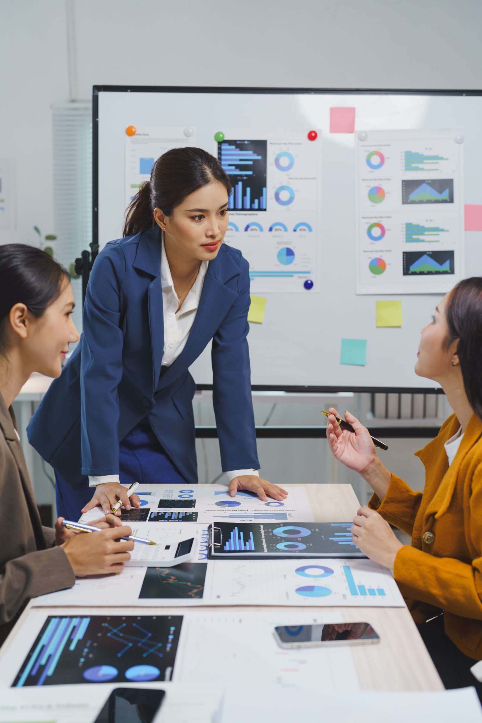 Asian businesswomen are analyzing financial reports, using data from charts and graphs displayed on a whiteboard and printed documents, working together to develop a successful marketing strategy
